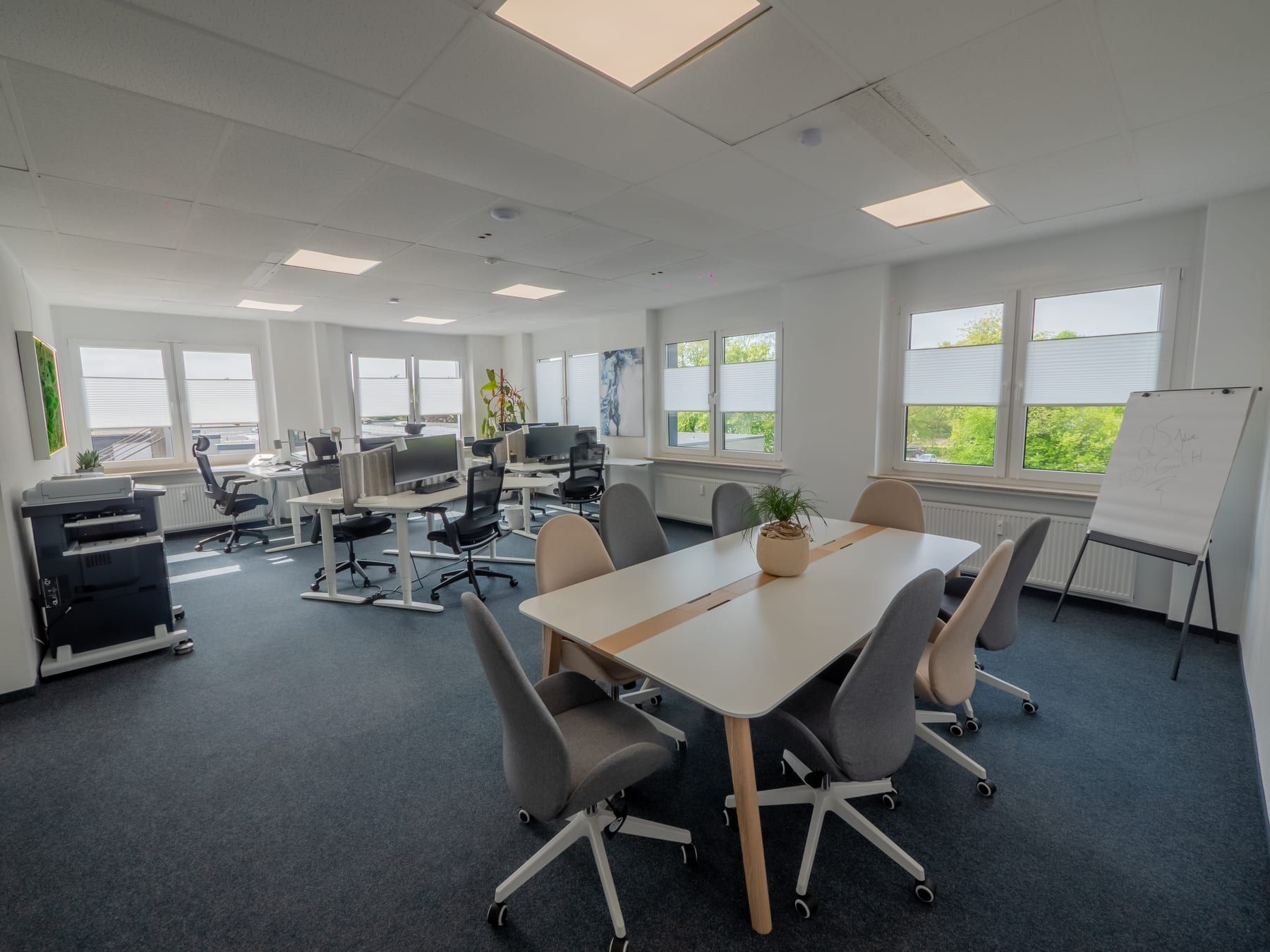 Open-plan office with a meeting table, flipchart and plotter, window front facing greenery
